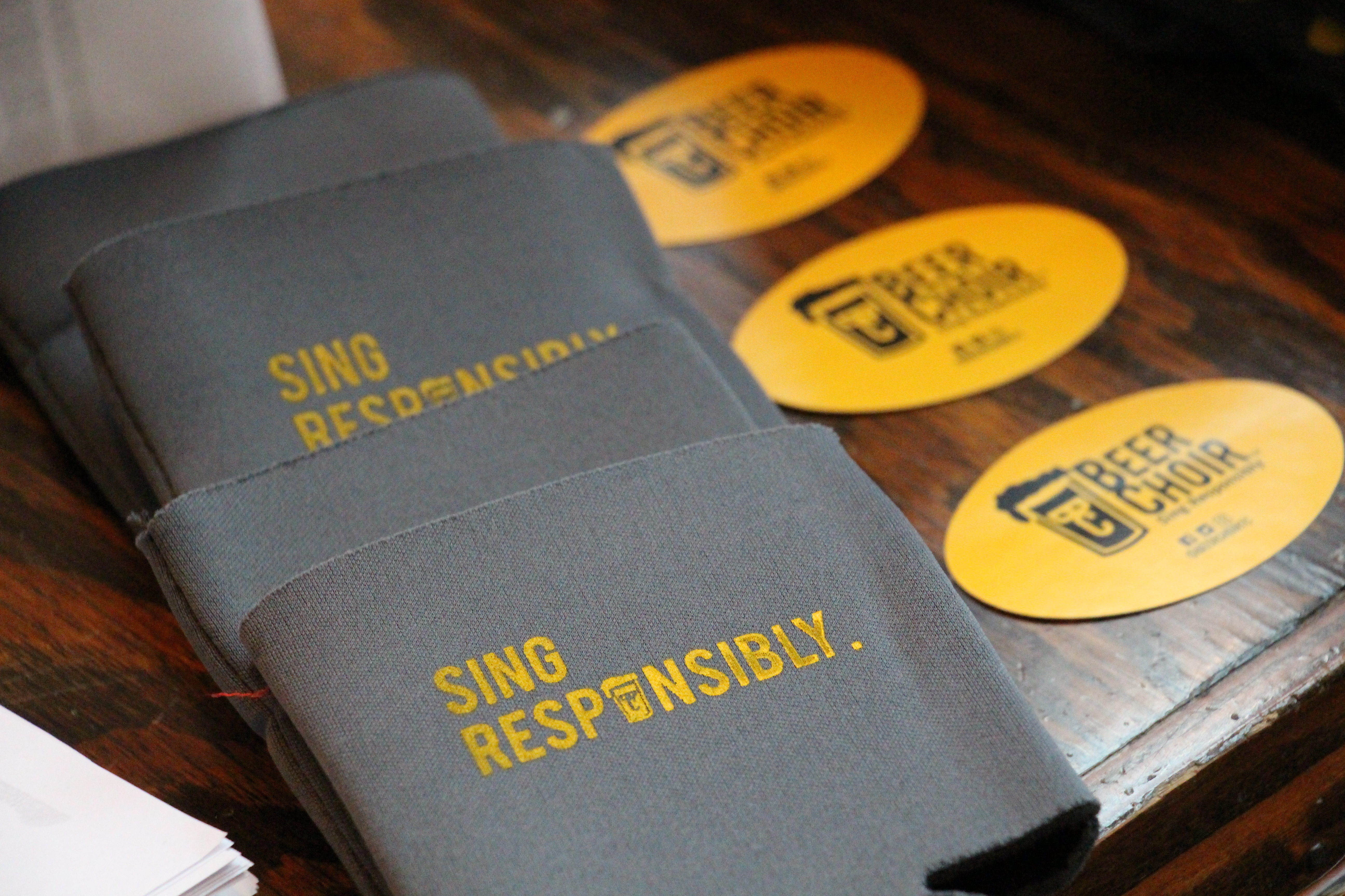 A close-up view of branded "Beer Choir" promotional items displayed on a dark wood surface at Price Creek Event Center. The image features several gray fabric beverage koozies printed with the phrase "SING RESPONSIBLY" in yellow lettering, alongside bright yellow oval stickers showcasing the Beer Choir logo. These items reflect the engaging atmosphere of our dinner theater and community events. Our full-service bar and flexible event space provide an ideal backdrop for unique gatherings, corporate retreats, and celebrations in the heart of Eastern Iowa.