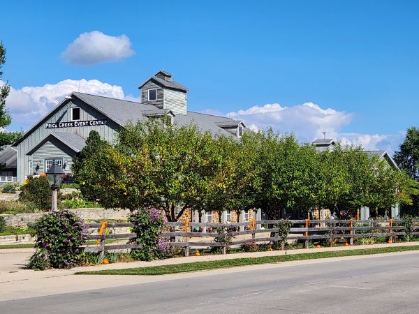 A clear, sunny day view of the Price Creek Event Center exterior. The image captures our classic, rustic barn-style venue building framed by a row of lush green trees and a wooden fence along the roadside. It showcases the inviting and professional curb appeal of our property, highlighting the spacious grounds and the prominent signage of the event center under a bright blue sky. This photo is perfect for introducing our venue as a welcoming location for weddings, corporate gatherings, and community events.