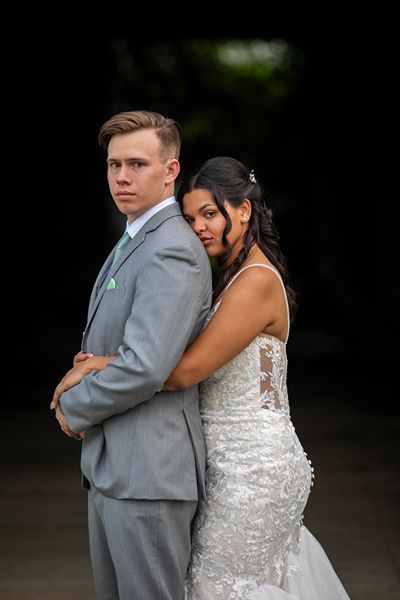 Bride and groom embrace outdoors in front of a rustic, shaded tunnel. The bride wears a fitted lace gown with a long train and the groom is in a light grey suit. A bouquet of white and magenta flowers rests on the grass beside them, highlighting a romantic wedding moment at Price Creek Event Center.