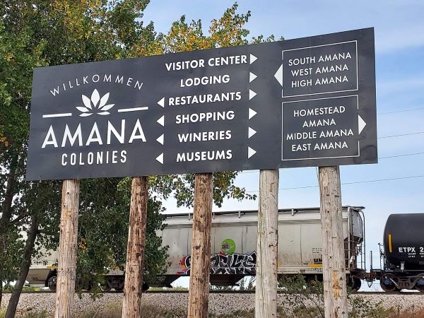 This image features the prominent "Willkommen" directional sign for the Amana Colonies. Mounted on wooden posts, the sign acts as a navigational guide for tourists, pointing toward the Visitor Center, local lodging, restaurants, shopping, wineries, and museums across the seven historic villages. In the background, a freight train passes by, highlighting the blend of our historic rural setting with active industrial transportation. This sign serves as the primary welcoming point for guests planning their itinerary to explore our unique cultural heritage and local attractions.