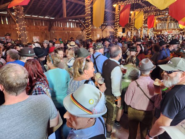 A high-angle shot capturing a dense, lively crowd inside our rustic barn during Oktoberfest. Guests are dressed in traditional German attire, including hats and lederhosen, holding beer glasses while socializing. The venue is decorated with vibrant, hanging pennant flags and warm string lighting, showcasing the energetic, packed atmosphere of our event.