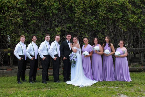 A professional wedding portrait featuring the bride and groom standing center, surrounded by their bridal party in front of a rustic, ivy-covered wall. The groomsmen are dressed in white shirts with suspenders and black trousers, while the bridesmaids wear matching lavender floor-length gowns. This image highlights the coordinated color palette and the natural, outdoor backdrop of our venue, perfect for showcasing the atmosphere of a classic wedding ceremony.